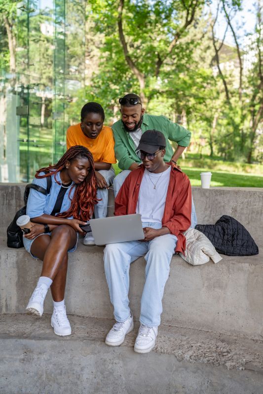 Excited black friends watch movie or make video call on laptop outdoor. Four African students focused on screen, laughing, engaged in social media, internet surfing, sharing idea, preparing for exam